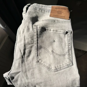 Ljusgrå jeans från Jack & Jones - Säljer ett par ljusgrå jeans från Jack & Jones med klassisk femficksdesign och diskreta slitningar på benen. Jeansen har rak passform och är perfekta för en avslappnad stil. Märkespatch i läder baktill i midjan.