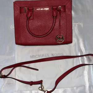 Röd handväska från Michael Kors.Äkta - Snygg röd handväska från Michael Kors med guldfärgade detaljer och avtagbar axelrem. Väskan har ett klassiskt MK-hänge och stilren design. Perfekt accessoar för att lyfta din outfit.Skydds påsen kommer med.