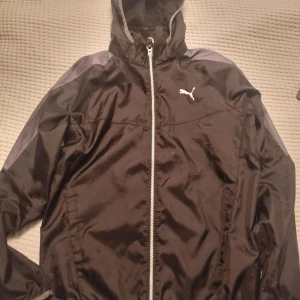 Svart vindjacka från Puma - Svart vindjacka från Puma med dragkedja och huva. Jackan har långa ärmar och en vit Puma-logga på bröstet. Storlek 164 enligt lapp, passar även xs herr.