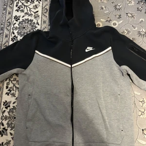 Nike tech fleece  - Säljer en grå och svart Nike tech fleece med dragkedja och huva. Tröjan har en vit randdetalj över bröstet och sidofickor. Perfekt för en sportig look eller chill dagar. Två klorin fläckar som syns på baksidan och dragkedjan går inte att stängas för att saken är borta på den men går att bara lägga till en ny. För stor på mig, priset kan dieksuteras!