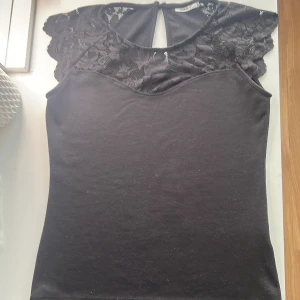 Svart topp med spetsdetaljer - Snygg svart topp från Only med spetsdetaljer på axlarna och övre delen. Toppen har en rund halsringning och en liten öppning bak. Perfekt för en stilren look. Hundra kr +frakt priset kan diskuteras