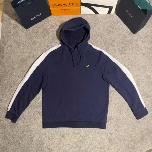 Mörkblå hoodie från Lyle & Scott - Säljer en snygg mörkblå hoodie från Lyle & Scott med vita ränder längs ärmarna och en gul logga på bröstet. Perfekt för en avslappnad stil. Den har en klassisk huva med dragsko och en stor ficka framtill. Skriv vid funderingar eller frågor:)