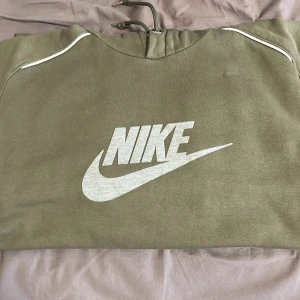 Grön hoodie från Nike - Säljer en grön hoodie från Nike med klassisk logga framtill. Hoodien har en justerbar huva med snören och ribbade muddar vid ärmslut och nederkant. Perfekt för en avslappnad stil.