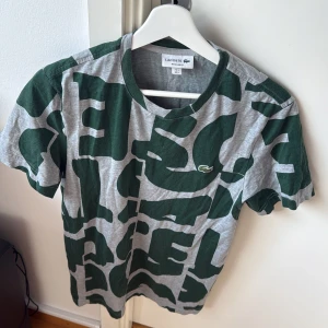 Grå och grön t-shirt från Lacoste - Säljer en grå och grön t-shirt från Lacoste med ett unikt mönster. T-shirten har korta ärmar och är perfekt för en avslappnad stil. Den ikoniska Lacoste-loggan syns på bröstet. Passar bra till jeans eller shorts.