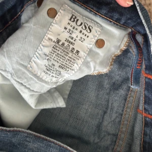 Blå jeans från Hugo Boss - Snygga blå jeans från Hugo Boss med klassisk femficksdesign och en lätt slitning på ena fickan. Jeansen har en knappgylf och orangea sömmar som ger en cool kontrast. Perfekta för en avslappnad stil.