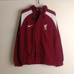 Liverpool jacka - Säljer en vinröd vindjacka från Nike med vita detaljer och L.F.C-logga på bröstet. Jackan har en dragkedja framtill och justerbar huva med snörning. Perfekt för sportiga tillfällen eller som en stilren vardagsjacka.