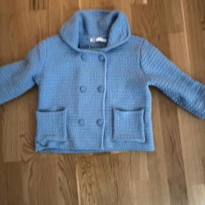 Blå stickad cardigan med knappar - En blå kappa i cardiganmatrial med dubbelknäppning och stora knappar. Jackan har en hög krage och två framfickor.