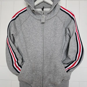 Grå hoodie från Key Largo - Säljer en grå hoodie från Key Largo med dragkedja och huva. Den har långa ärmar med ränder i rött, vitt och svart längs sidorna. Perfekt för en avslappnad stil.