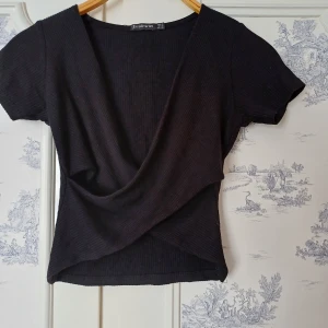 Svart ribbad omlottopp från Stradivarius - Säljer en stilren svart ribbad omlottopp från Stradivarius. Toppen har en v-ringad design och korta ärmar, perfekt för en trendig look. Den är tillverkad i ett mjukt och bekvämt material som sitter snyggt på kroppen.