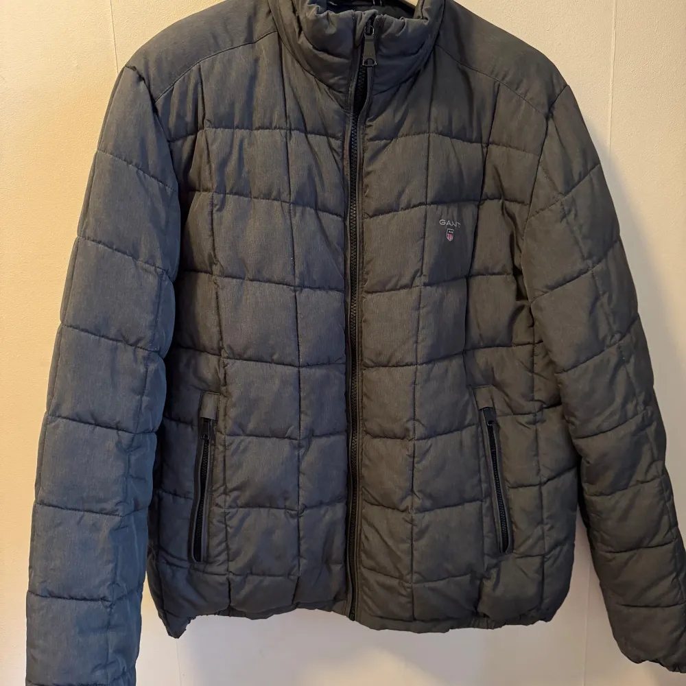 Säljer en stilren grå quiltad jacka från GANT. Jackan har en dragkedja framtill och är långärmad. Perfekt för kyligare dagar med sin pufferdesign som håller värmen. Den har en diskret logga på bröstet och praktiska fickor med dragkedja.. Takit.
