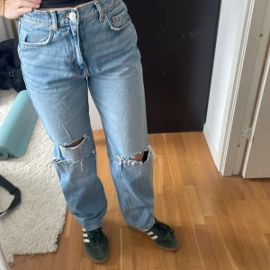 Blå jeans från Gina Tricot - Snygga blå jeans från Gina Tricot med hög midja och slitna detaljer på knäna. De har en klassisk femficksdesign och en knappgylf. Perfekta för en avslappnad stil.
