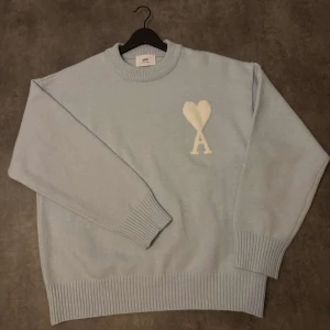 Ljusblå stickad tröja från Ami - Säljer en stilren ljusblå stickad tröja från Ami med ett vitt hjärtmönster och bokstaven A på bröstet. Tröjan har långa ärmar och ribbade muddar vid ärmslut och nederkant. Perfekt för en avslappnad och trendig look. Storleken är M men passar även som L.