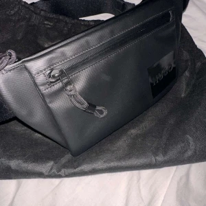 Svart axelväska från Hugo - Snygg svart axelväska från Hugo med dragkedja framtill. Väskan har en stilren design och justerbar rem för bekväm användning. Perfekt för att bära med sig det viktigaste på ett smidigt sätt. vi kan diskutera om priset