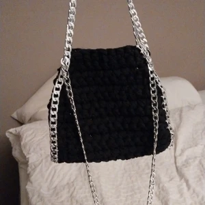 Svart virkad väska med kedjedetaljer - Snygg svart stickad väska med coola silverfärgade kedjor. Perfekt för att ge din outfit en edgy touch. Väskan har en unik textur och är både stilren och praktisk.