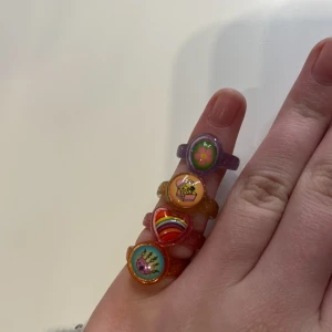 Färgglada ringar med motiv - Säljer ett set med fyra färgglada ringar i plast. Varje ring har ett unikt motiv: en hand med färg, ett hjärta med regnbåge, en cupcake och en blomma. Ringarna är i olika färger som orange, rosa, guld och lila.