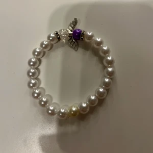 Pärlarmband med ängelberlock - Charmigt armband med vita pärlor och en unik ängelberlock i silver. Armbandet har även en lila och en klar pärla som ger en fin kontrast. Perfekt för att ge en personlig touch till din stil.