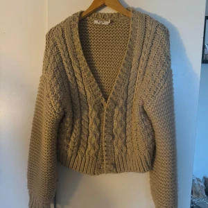 Beige stickad kofta från NA-KD - Säljer en mysig beige stickad kofta från NA-KD. Koftan har en v-ringad design med knappar framtill och ett fint flätmönster. Perfekt för kyliga dagar!