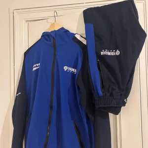 Blå och svarta träningsbyxor från Yamaha - Snygga träningsbyxor från Yamaha i blått och svart med logotyp på benet. Byxorna har en elastisk midja och är perfekta för sportiga aktiviteter. De har en modern design med kontrasterande färgdetaljer.