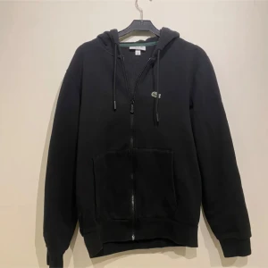 Svart hoodie från Lacoste - Säljer en klassisk svart hoodie från Lacoste med dragkedja och huva. Tröjan har långa ärmar. Perfekt för en avslappnad stil. Nypris 1400 kr