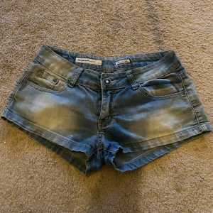 Lågmidjade shorts - Snygga blå jeansshorts från Miss Me med slitna detaljer och låg midja. Perfekta för sommardagar med en avslappnad stil. Shortsen har klassiska fem fickor och knappgylf.