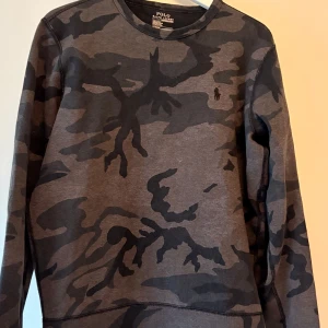 Camouflage tröja från Polo Ralph Lauren - Säljer en snygg camouflage tröja från Polo Ralph Lauren. Tröjan har en klassisk rund hals och är perfekt för en avslappnad stil. Den är i en mörk färgskala med det ikoniska Polo-logotypen på bröstet.