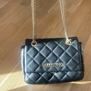En svart axelväska från Valentino - Snygg svart axelväska från Valentino med guld detaljer. använd få gånger då jag har silver på mig och det passar inte så bra med en väska med guld detaljer❣️
