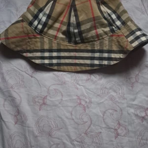 Burberry bucket hat - Snygg bucket hat med klassiskt rutigt mönster i beige, svart och rött. Perfekt för att ge din outfit en trendig touch. Passar bra till både vardag och speciella tillfällen.