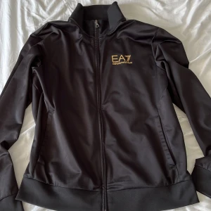 Emporio Armani zip Hoodie svart - Svart zip hoodie med guldigt EA7 märke på vänster bröst, använder inte längre, köpt för 1000kr på johnells. Storlek L passar M bra