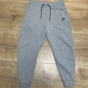 Nike Tech Byxor - Säljer mina gråa Nike Tech Byxor i storlek M då dom inte passar mig längre. Dom är jätte lite trasiga vid högra delen som man kan se på bild 3 om man tittar noggrant men det är knappt synligt, annars i helt nytt skick. 