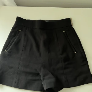 Shorts - Svarta shorts med gulddetaljer, finns en dragkedja på sidan samt knapp. Finns även två fickor i fram och två falska fickor i bak. 