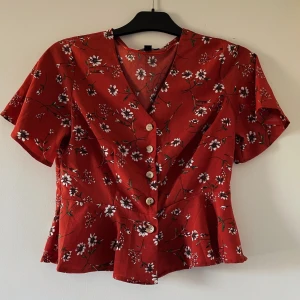 Blommig topp - T-shirt topp i skjortmaterial. Tight om bröstet men utsväng runt midjan. Blommigt material. Knappt använd, praktiskt taget nyskick. 