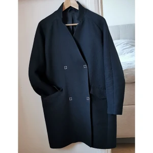 Lätt vadderad vårkappa J.Lindeberg - Dubbelknäppt kappa i fint skick. Boxig passform. Tryckknappar. Fickor med lock. Medellång 90 cm, slutar ovanför knät. Kort slits bak, 20 cm. Speciellt och oregelbundet tyg. Yttertyg: 80% bomull, 10% polyester, 10% polyamid. Foder: 95% bomull. 5% elastan. 