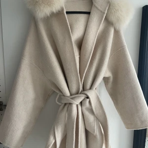 Beige kappa med äkta pälskrage  - En oanvänd beige kappa med en äkta pälskrage. Går precis ovanför knäna. 