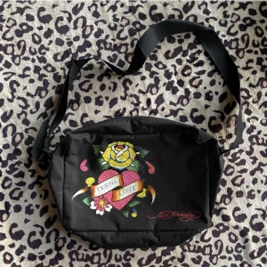 ed hardy väska - Säljer min jättefina Ed Hardy väska då den inte kommer till anändning. Den får plats med dator osv.  Några rhinestones har trillat av men det är inte något som man märker av💋 skriv om du vill ha fler  bilder!