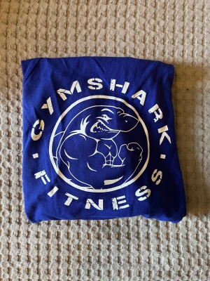 Gymshark top - Top från Gymshark i jättefin blå färg. Med tryck på ryggen och hål för tummarna.  Aldrig använd, endast provad.  Tyvärr lite för stor för mig.  Storlek M  Finns i Vallentuna! Köpare står för frakt! 