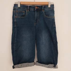Blå shorts i fejk jeans material, med stretch material. Har två bakfickor och är precis ovanför knäna. Bra skick