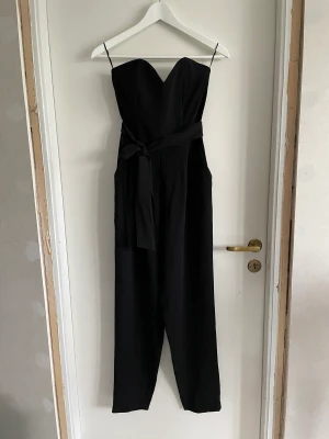 Ärmlös byxdress - Jumpsuit / byxdress med skärp i midjan. Stängs med dragkedja på sidan. Gummerat/silikon i kant över byst för bättre fäste.