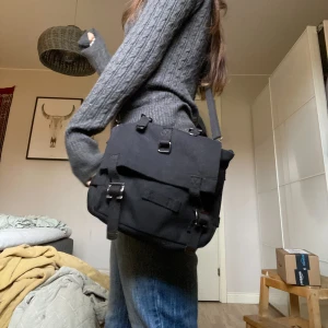 Svart väska med detaljer - Svart axelväska med många detaljer. Väskan är i tyg och har en justerbart band. Den är använd men i gott skick utan skador eller grova defekter. Skriv gärna för fler bilder eller andra frågor!🖤🐦‍⬛