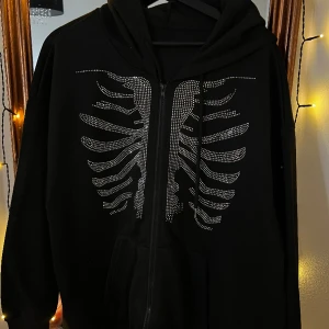 Cool Hoodie med dragkedja  - Svart hoodie oversizetill försäljning! Den är tillverkad av mjuk och elastisk bomull och har en snygg dragkedja framtill. Det unika med denna hoodie är att den har en vacker revbensdetalj gjord av glittrande kristall