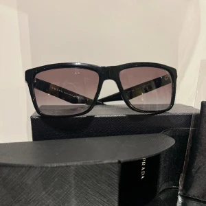Prada solglasögon  - Solglasögon från Prada. Svarta med en mörklila ton på glaset. Använt väldigt sparsamt under en sommar. Topp skick med original förpackning samt kvitto. Ordinarie pris 2550kr.