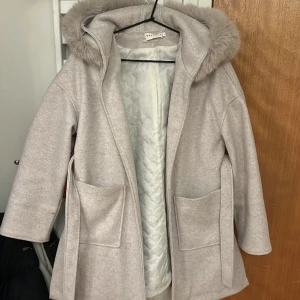Wool Blend Coat - En kort kappa med skärp och stor luva omgiven av päls. Fuskpälskragenr avtagbar. Kappan är enfärgad i beige med två fickor, en på vardera sida. Kappan är i storlek S. Passformen är liten. Kappan har inga fläckar, den är som nyköpt <3