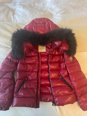 Monclerjacka - Världens finaste jacka! Vinröd och med en superfin krage och märke samt ficka på vänster arm. ”Historien” på insidan är lös samt är hängöglan sönder, i övrigt har den inga defekter. Storlek 152 men skulle säga  större i storlek. 