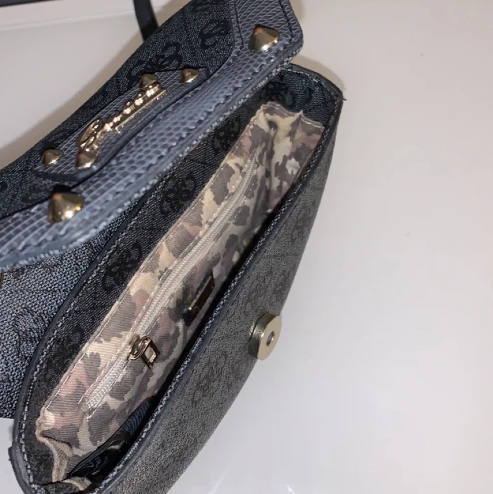 Cool lite mindre mörkgrå Guess väska! Den har fina detaljer som nitar och leopardmönster inuti, samt en innerficka. . Väskor.
