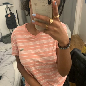 Rosa och vit randig Lacoste T-shirt - En Lacoste T-shirt som inte kommit mycket till användning vilket är varför jag säljer den! Skulle säga att den passar både XS, S & M☺️