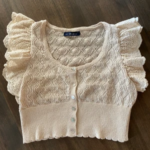 Söt virkat topp i cremevit - Ur gullig virkad topp i storlek S. Den har jätte fina axel detaljer och pärlemo knappar, den är cropped och slutar ovan naveln.