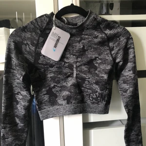 GymShark Grå Camo Topp - Helt oanvänd träningstopp beställd från Gymshark. Croppad passform, ej genomskinligt tyg med dekorativa hål i rygg samt för tummarna. Säljer då den aldrig kommit till användning. Topp (XS) 150kr, Leggings (S) 300kr, Hela Setet 400kr. Kan mötas i Uppsala eller skicka (frakt 45kr + 20kr kostnadstillägg om SafePay önskas). Fler plagg från Gymshark i min profil.