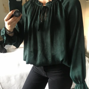 Mörkgrön blus - Mörkgrön luftig blus med volanger i ärmsluten och halsringen, knytning vid halsen och resår vid höften