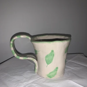 Handgjord kopp med gröna löv - Koppen är ofärgad med ljusgröna löv på samt gröna ränder på örat. 