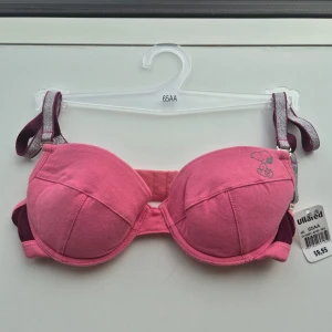 Rosa Snoopy-bh 65AA - Söt rosa (rosaröd) bygel-bh från 00-talet med uttagbara kuddar, justerbara och avtagbara axelband i silver. Stängs med hyskor bak. Snobben-tryck på ena kupan. Perfekt för dig som vill ha en färgglad och bekväm bh. Orginallappen är kvar, denna är oanvänd med kommer från ett restlager. Äkta Y2K. Finns även i storlek 75A och 75B, bara skriv till mig. Bundlerabatt på alla mina varor.