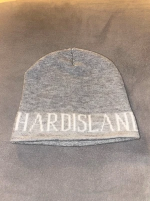 Grå mössa med Hard Island-tryck - Snygg grå mössa med vit text där det står 'HARD ISLAND' längs kanten. Klassisk modell i mjukt stickat material som passar perfekt när det är kallt ute. Enkel och stilren design som funkar till de flesta outfits.